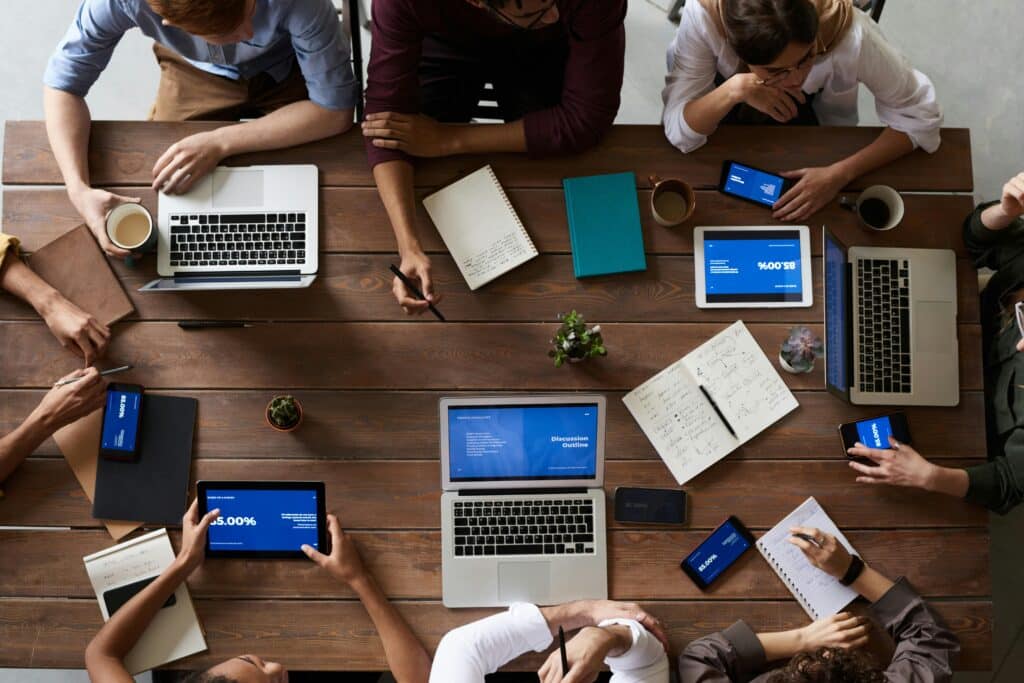 Eine Gruppe von Personen sitzt rundherum um einen Tisch in einer Besprechung mit ihren Laptops und Tablets.